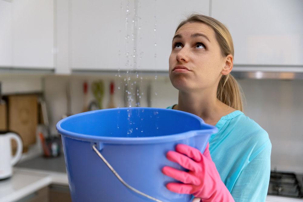 Woman holding a blue bucket under water leaking from the ceiling in a kitchen
