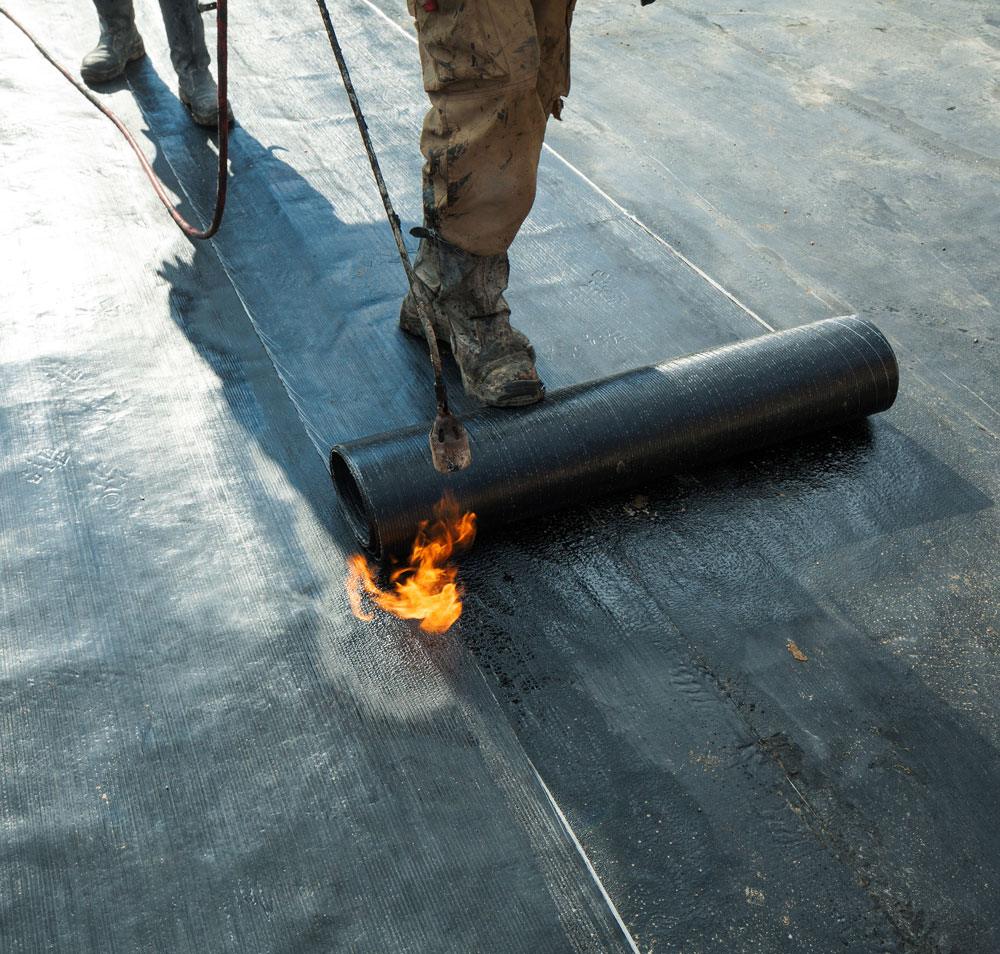 Worker using a torch to heat a roll of black roofing membrane on a flat roof