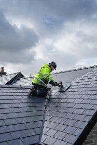 Worker in high-visibility jacket kneeling on a slate roof under cloudy skies