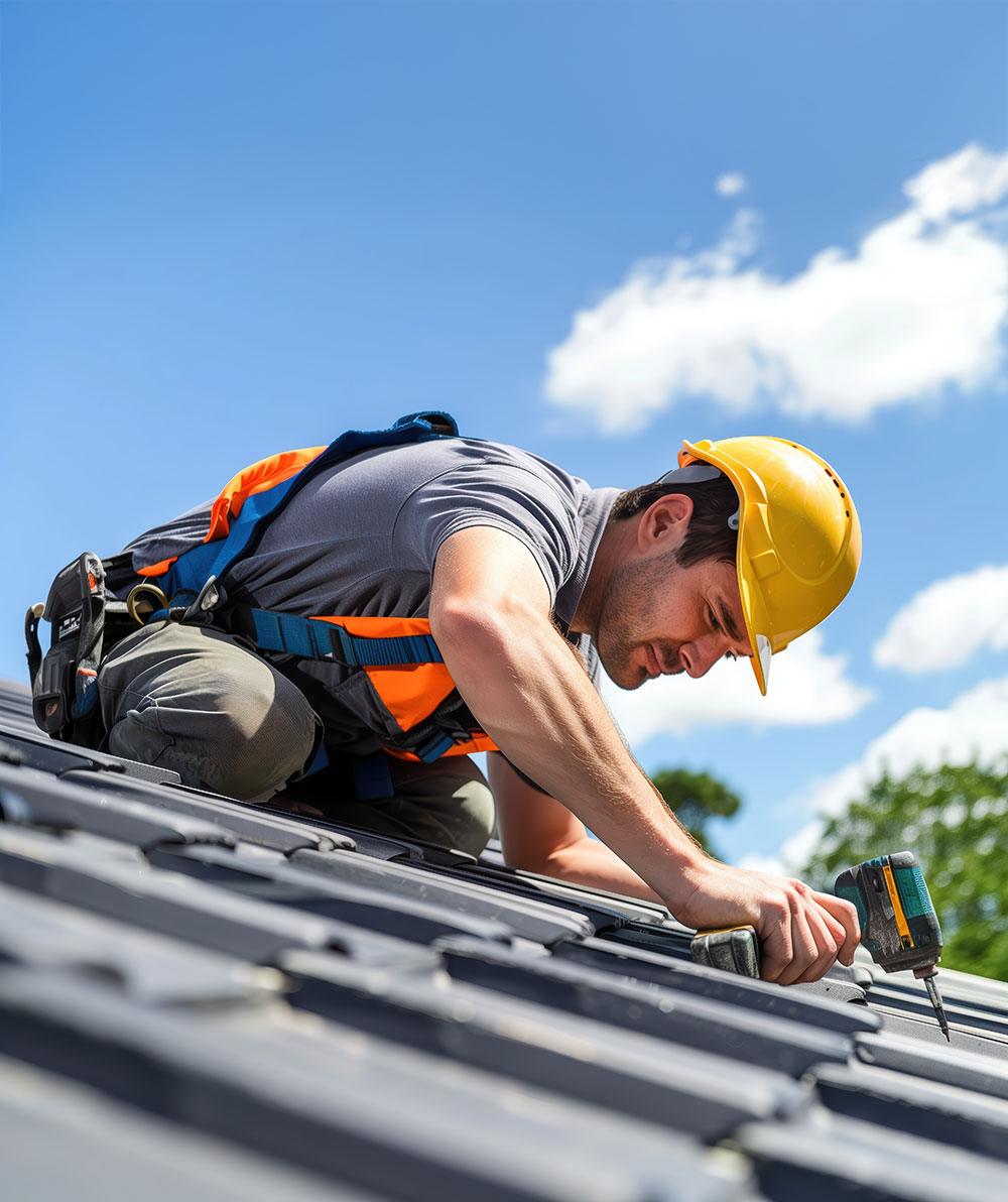 Worker in a yellow hard hat using a drill on a tiled roof under a blue sky